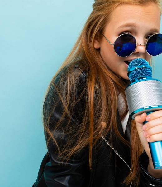 Nice girl in black jacket, blue sunglasses sings songs with blue microphone isolated on blue background Teenager in black jacket, blue sunglasses sings songs with blue microphone isolated on blue background